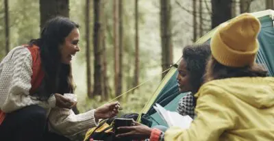 Three adult women on a camping trip make coffee near a tent
