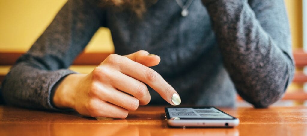 A phone sits on a table. A woman leans over it, scrolling with one finger.