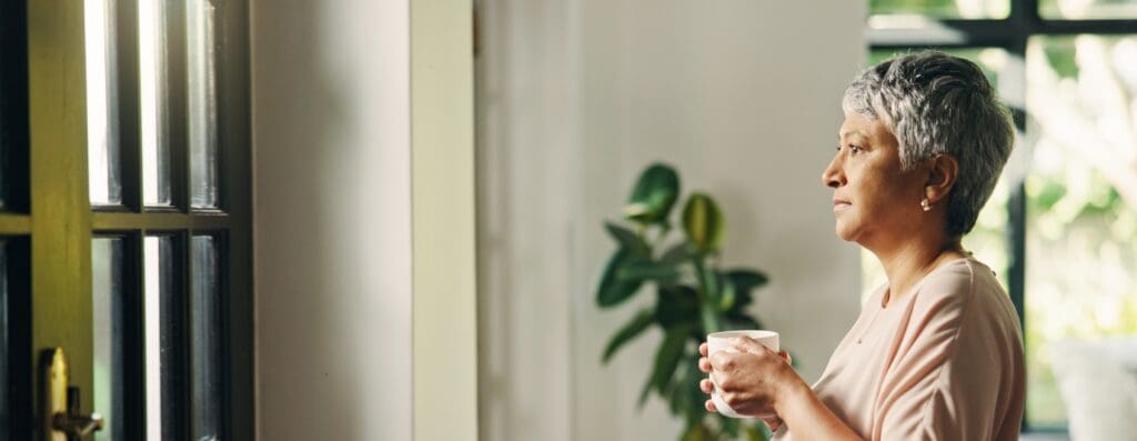An older woman holding a cup of coffee stares out a window with a blank expression