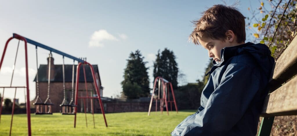 A boy sits alone on a bench at a park looking down
