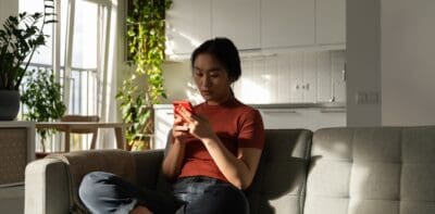 A woman sits on her couch looking at her phone and thinking seriously