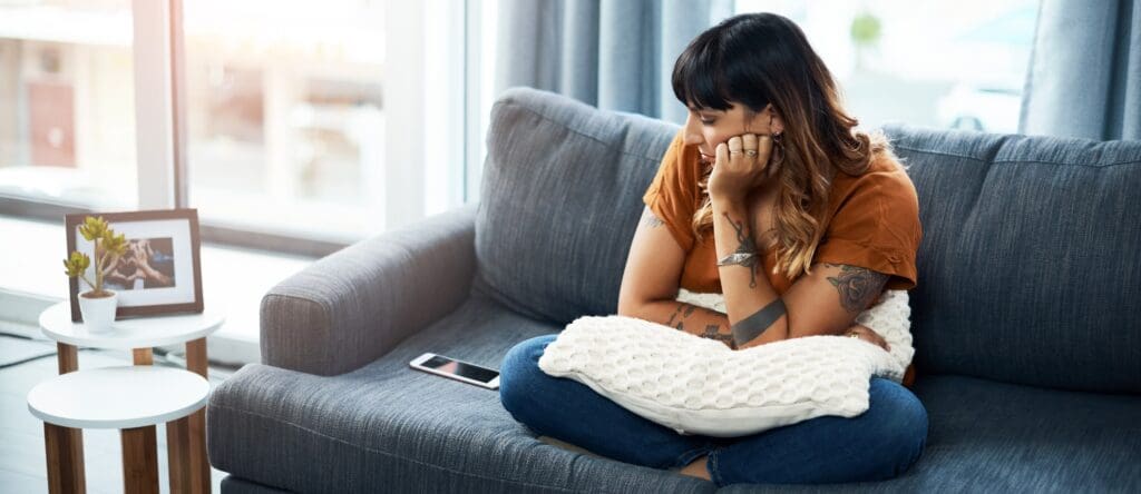 A woman sits on her couch looking at her phone resting beside her, thoughtful and sad