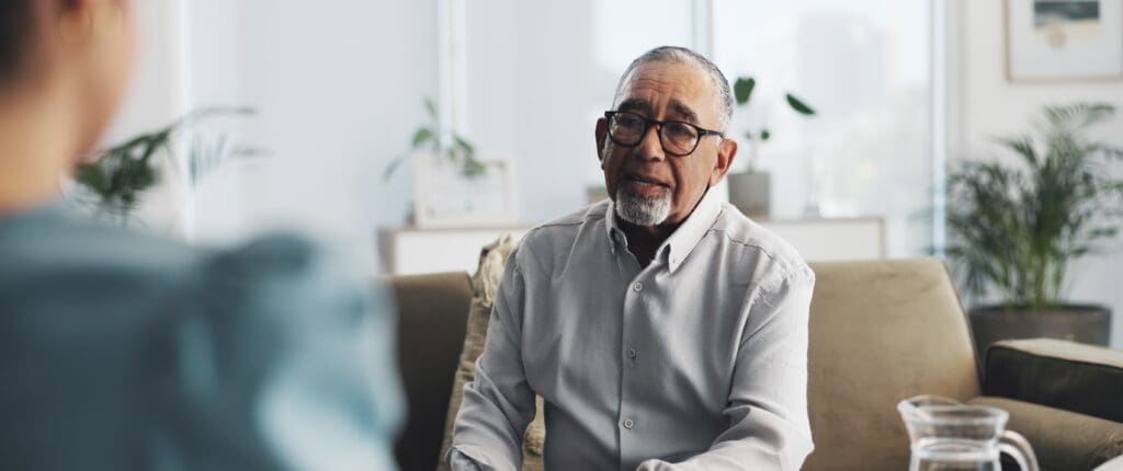 An older gentleman with a thoughtful expression sits on a couch talking to a female therapist
