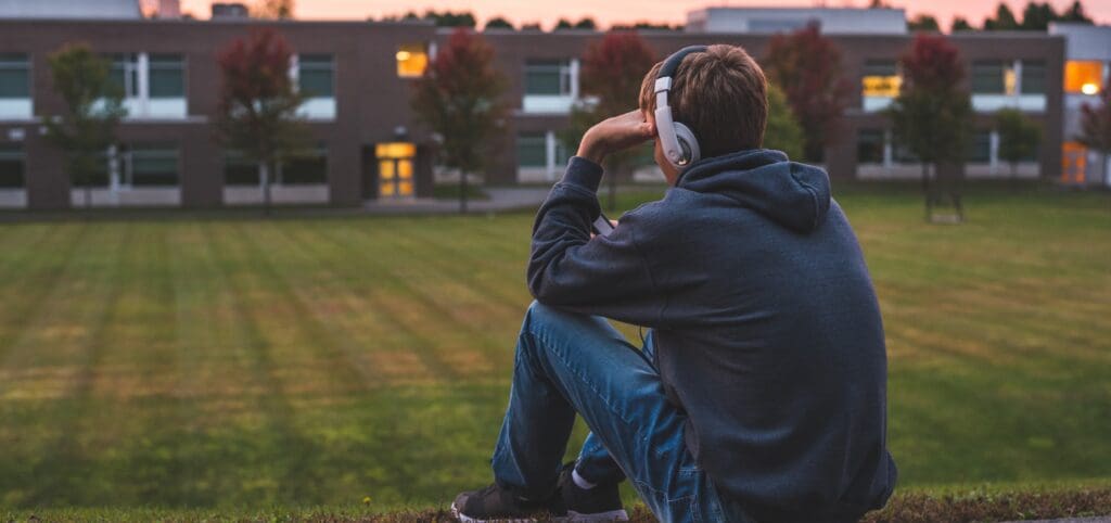 A teenage boy sits on a hill overlooking his school listening to music with his hand pressed to his head, worried